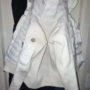 Vit jacka från Moncler med quiltade detaljer - Säljer en vit Moncler-jacka med quiltade partier och ribbstickade ärmar. Jackan har dragkedja, huva och klassisk Moncler-logga på ärmen. Insidan har ett färgglatt serietidningsmärke. Perfekt för dig som vill ha en stilren och exklusiv look, pris kan också diskuteras 