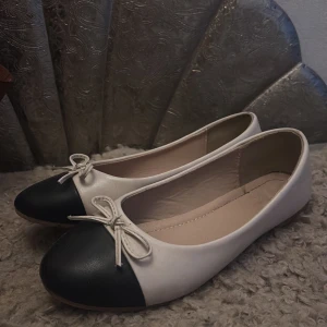 Svartvita ballerinaskor med rosett - Säljer ett par klassiska ballerinaskor i vitt med svart tå och söt rosettdetalj framtill. Skorna har rund tå och platt sula, perfekta för en stilren look. Passar till många olika outfits och är superenkla att matcha.