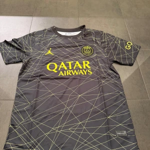 PSG fotbollströja Jordan x Qatar Airways - Säljer en svart PSG fotbollströja med gula detaljer och tryck från Jordan och Qatar Airways. Tröjan har korta ärmar, geometriskt mönster i gult och Paris Saint-Germain-logga på bröstet. Perfekt för fotbollsfans som vill sticka ut på planen.