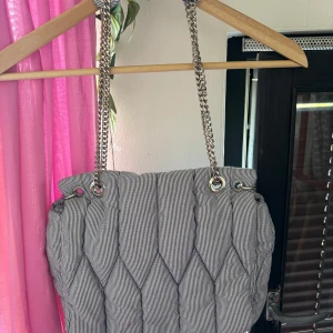 Randig axelväska med kedjerem - Säljer en snygg axelväska med smala svarta och vita ränder. Väskan har quiltad design och en silverfärgad kedja som axelrem. Perfekt för dig som vill ha en stilren accessoar med det lilla extra.