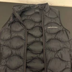 Svart dunväst från Peak Performance - Säljer en svart dunväst från Peak Performance med diskret logga på bröstet. Västen har quiltad design och dragkedja framtill. Perfekt för lager-på-lager och håller dig varm under kyliga dagar.