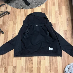 Svart vindjacka med huva från Lacoste - Säljer en svart vindjacka från Lacoste med huva och dragkedja framtill. Jackan har en minimalistisk design med Lacoste-loggan på bröstet och två sidofickor. Perfekt för blåsiga dagar.