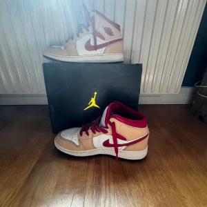 Nike Air Jordan 1 Mid Beige/Röd - Säljer ett par Nike Air Jordan 1 Mid i beige och rött med vita paneler och röda detaljer. Skorna har klassisk hög siluett, röd swoosh och snörning. Perfekta för dig som gillar streetwear och sneakers med ikonisk design.
