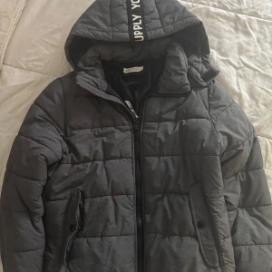 Grå quiltad pufferjacka med huva - Säljer en grå pufferjacka med huva och dragkedja framtill. Jackan har quiltad design och två fickor med knappar. Perfekt för kyligare dagar och har en sportig look med textdetalj på huvan. Till barn 8-9 år.