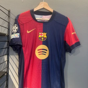 FC Barcelona hemmatröja 2023/24 Lamine Yamal #19 - Säljer en officiell FC Barcelona hemmatröja från säsongen 2023/24 med Lamine Yamal och nummer 19 på ryggen. Tröjan är röd och blå med guldiga detaljer, klubbmärke på bröstet och Champions League-märke på ärmen. Spotify-logga på framsidan. Helt ny med tagg en replika