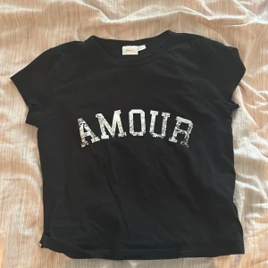 Svart t-shirt med text från Gina Tricot - Svart t-shirt från Gina Tricot med vit text 'AMOUR' framtill. Klassisk rund halsringning och korta ärmar. Perfekt för en avslappnad och cool stil.