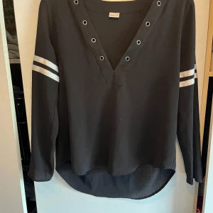 Svart långärmad tröja med vita ränder från Gina Tricot - Svart långärmad tröja från Gina Tricot med två vita ränder på varje ärm. Tröjan har v-ringning med metallöljetter och en något längre bakdel. Perfekt för en avslappnad och sportig stil. Är storlek xs men skulle mer säga S kanske till och med M. Tyvärr är snören till den borta men går att ersätta med vanliga svarta snören 