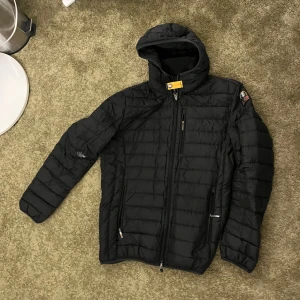 Svart dunjacka från Parajumpers - Säljer en svart dunjacka från Parajumpers med huva och dragkedja. Jackan har quiltad design, flera fickor med dragkedja och en gul detalj vid kragen. Perfekt för kyliga dagar. OBS: finns ett hål vid högra arm som går att fixa hos skräddaren för 300kr säljes så.Storlek L passar som M oxå.Hör av er för fler frågor!!
