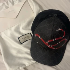 Svart keps från Gucci med orm-motiv - Snygg svart keps från Gucci med klassiskt GG-mönster och en röd-vit orm broderad på framsidan. Kepsen har böjd skärm och mesh på baksidan för extra ventilation. Perfekt accessoar för att lyfta din stil.