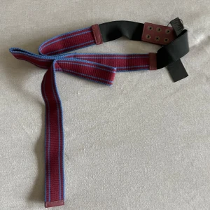 Randigt bälte från Maje - Snyggt bälte från Maje i vinrött med blå ränder och svarta detaljer. Bältet har läderdetaljer och metallöljetter. Perfekt accessoar för att lyfta din outfit med en färgklick och cool design.