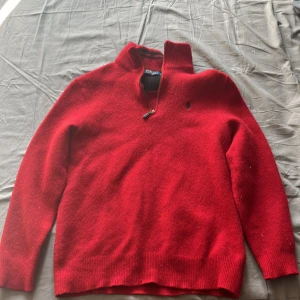 Röd ulltröja med dragkedja från Polo Ralph Lauren - Säljer en klassisk röd tröja från Polo Ralph Lauren i ull med halv dragkedja och broderad logga på bröstet. Tröjan har hög krage och långa ärmar, perfekt för kyliga dagar.