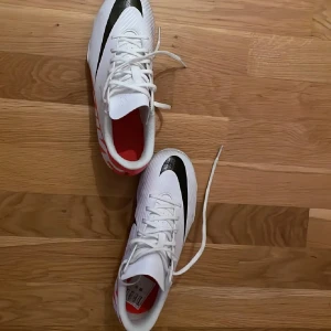 Vita Nike fotbollsskor med svart swoosh - Säljer ett par vita Nike fotbollsskor med svart swoosh och röda detaljer. Skorna har snörning och dobbar för spel på gräs/konstgräs. Perfekta för dig som vill ha snygga och funktionella skor på planen. Använts 3 gånger och säljs eftersom dottern valt att sluta med fotbollen.