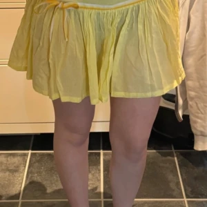 Gul kortkjol med rosett - Söt gul kortkjol med volang och knytband i midjan. Kjolen har en luftig och lätt känsla, perfekt för varma dagar. Den har en liten rosett framtill och är tillverkad i ett tunt, mjukt tyg.