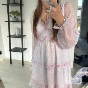 Ljusrosa blommig klänning med volang - Supersöt ljusrosa kortklänning med småblommigt mönster och volangdetaljer från Vero Moda. Klänningen har långärmade, transparenta ärmar och v ringning. Perfekt för sommaren! 