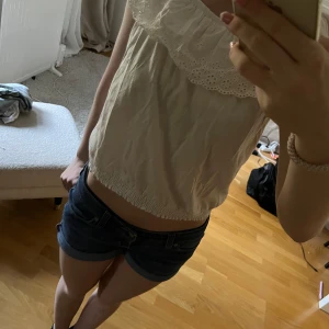 Vit blus med broderad volang - Söt vit blus med bred broderad volang runt axlarna och resår nertill. Blusen är ärmlös och har en romantisk känsla tack vare spetsdetaljerna på volangen. Perfekt för varma dagar. Denna topp är i storlek 146-152