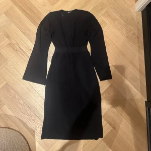 Svart långärmad klänning från Gina  - Snygg svart klänning från Gina med lång ärm och v-ringad hals. Klänningen har en markerad midja och är figurnära med en elegant, enkel design. Perfekt för dig som gillar stilrena plagg med lite edge.