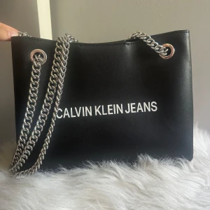Svart axelväska Calvin Klein Jeans - Snygg svart axelväska från Calvin Klein Jeans med silverfärgade kedjehandtag och logotyp i vitt på framsidan. Väskan har en stilren, rektangulär form och är tillverkad i skinnimitation. Säljer pga att den nt kommer till användning! Pris kan diskuteras 