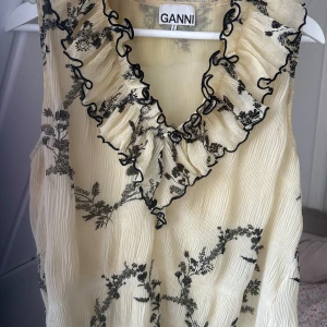 Blommig volangblus från Ganni - Supersöt blus från Ganni i ljusgul nyans med svarta blommiga mönster. Blusen är ärmlös och har en djup v-ringning med volangkant och svarta broderade detaljer. Materialet är lätt plisserat och väldigt fint. 💕💛💝