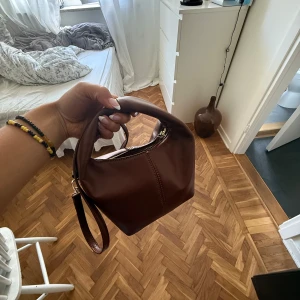 Brun handväska i skinn med axelrem - Säljer en snygg brun handväska i skinn med mjuk form och guldfärgade detaljer. Väskan har både ett kort handtag och en längre axelrem, samt dragkedja upptill. Perfekt storlek för det viktigaste och stilren look. Inga tecken på användning❤️