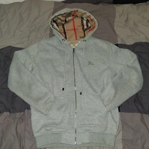 Burberry hoodie grå - Snygg grå hoodie från Burberry med klassiskt rutigt foder i huvan och broderad logga på bröstet. Tröjan har dragkedja framtill, snörning i huvan och två fickor. Storlek S