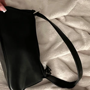 Svart väska  - Snygg svart axelväska i skinnimitation med minimalistisk design. Väskan har en bred axelrem, dragkedja upptill och en praktisk ytterficka med dragkedja. Perfekt storlek för det viktigaste och passar till många olika outfits.