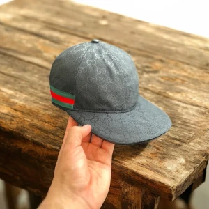 Gucci svart keps med GG-mönster - Svart keps från Gucci med diskret GG-monogram över hela ytan och klassiska gröna och röda ränder på sidan. Justerbar rem baktill med metallspänne. Tillverkad i bomull och polyester för en lyxig känsla och snygg passform.