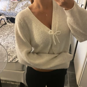 Vit kofta med pärlrosett - Superfin vit kofta med v-ringning och en söt rosett av pärlor framtill. Koftan är ribbstickad och har en lite loose passform med långa ärmar. Perfekt att slänga på sig för en chill och stilren look.