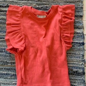 Koralröd volangtopp från Zara - Supersöt korallröd topp från Zara med korta ärmar och stora volangdetaljer längs axlarna. Toppen är ribbad och har en rund halsringning. Perfekt för dig som vill sticka ut med en färgglad och trendig look.