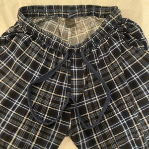 Rutig mjukisbyxa Ben Buxton XL - Blå och svart-rutiga mjukisbyxor från Ben Buxton i storlek XL. Byxorna har elastisk midja med snörning och är gjorda i mjukt bomullstyg. Perfekta för chill dagar hemma eller när du vill vara extra bekväm.