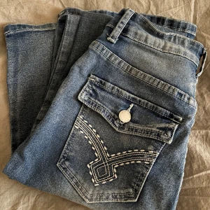 Blå jeans bootcut med broderad bakficka - blå jeans med kontrastsömmar och broderad bakficka. dragkedja och knapp framtill samt en fickdetalj med lock och knapp. 