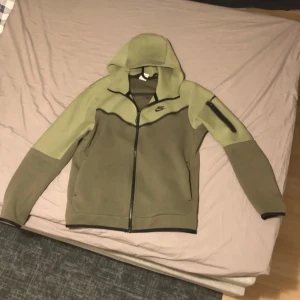 Nike hoodie jacka i olivgrönt - Nike hoodie jacka i två nyanser av olivgrönt med svart logga på bröstet. Jackan har hel dragkedja, huva och en praktisk ficka med dragkedja på ena ärmen. Tillverkad i mjukt syntetmaterial, perfekt för chill eller träning.