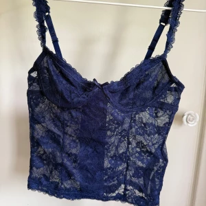 Mörkblå spetsig korsett-topp - Supersöt mörkblå korsett-topp i spets med blommigt mönster. Justerbara axelband med spetsdetaljer och liten rosett framtill. Perfekt för dig som vill ha en trendig och feminin look.