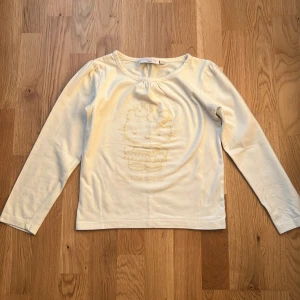 Hello Kitty långärmad vit tröja - Söt långärmad tröja från Hello Kitty . Framsidan har ett diskret Hello Kitty-relieftryck med rosett och text. Rund halsringning och rak passform, perfekt för dig som gillar gulliga detaljer och vill ha en chill look.