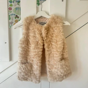 Beige pälsväst från Superior 1884 - Mysig och fluffig beige pälsväst från Superior 1884. Västen har en klassisk rak passform och är tillverkad i mjukt fuskpälsmaterial. Perfekt för att addera lite extra stil till din outfit.