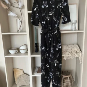 Svart blommig jumpsuit med bälte - Snygg svart jumpsuit med vita och grå blommor, knytbälte i midjan och korta ärmar. Perfekt för dig som gillar en stilren look med lite extra detaljer. Jumpsuiten har en avslappnad passform och är superenkel att matcha med accessoarer.