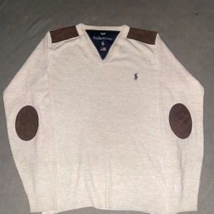 Beige v-ringad tröja Polo Ralph Lauren - Snygg beige v-ringad tröja från Polo Ralph Lauren med bruna mockadetaljer på axlar och armbågar. Broderad logga på bröstet. Tröjan är stickad och har långa ärmar, perfekt för lager-på-lager under höst och vinter.