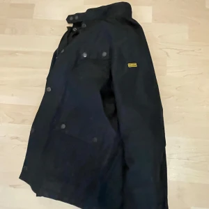 Svart Barbour International jacka storlek L barn jacka  - Snygg svart jacka från Barbour International med klassisk design och quiltat foder. Jackan har flera fickor med knappar, hög krage med spänne och dragkedja framtill. Perfekt för dig som vill ha en stilren och funktionell jacka med streetvibe.
