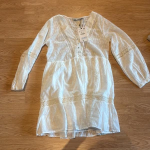 Vit kortklänning med spets från Zara - Supersöt vit kortklänning från Zara med diskret prickigt mönster och spetsdetaljer längs v-ringningen och ärmarna. Klänningen har långärmade ärmar, knappar framtill och volang vid nederkanten. Perfekt för sommarens alla tillfällen!