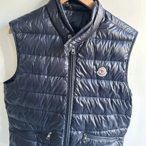 Moncler väst - Snygg svart dunväst från Moncler med glansig finish och klassisk logga på bröstet. Västen har hög krage, två dragkedjefickor och quiltad design. Perfekt för lager-på-lager och funkar året runt när du vill hålla stilen chill men ändå varm. Storlek 3 vilket motsvarar L. En liten skada på den men väldigt fin annars