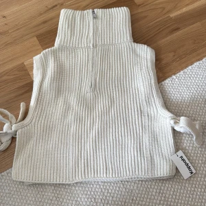 Vit stickad väst från KappAhl - Snygg ribbstickad väst i ljusvit från KappAhl med hög krage och half zip framtill. Västen har knytband i sidorna för extra stilpoäng och är perfekt att slänga över en t-shirt eller skjorta. Enkel, clean och superenkel att matcha. Prislappen kvar