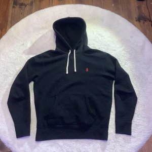 Svart hoodie från Polo Ralph Lauren - Svart hoodie från Polo Ralph Laurent strl S med klassisk röd broderad logga på bröstet. Tröjan har vit snörning vid huvan, ficka och långa ärmar. Mjuk bomullskvalitet som är perfekt för chill dagar. Finns även Tag-lappar
