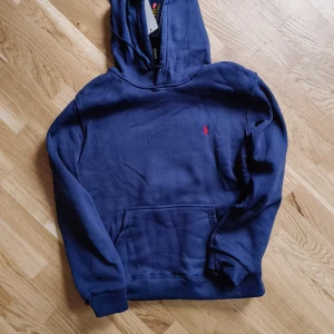 Marinblå hoodie från Polo Ralph Lauren - Snygg marinblå hoodie från Polo Ralph Lauren med klassisk röd logobrodyr på bröstet. Tröjan har huva, magficka och långa ärmar med muddar. Perfekt för chill dagar och enkel att matcha med jeans eller joggers.
