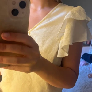 Ljusgul blus med volangärm - Supersöt ljusgul blus med v-ringning och volangdetaljer på ärmarna. Blusen har en lätt textur i tyget och är kortärmad, perfekt för dig som vill ha en feminin och fräsch look. Passar till jeans eller kjol för en chill stil.