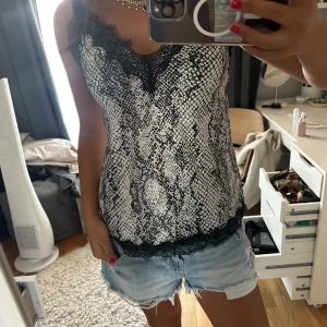 Svartvit linnetopp med spetsdetaljer - Säljer en snygg linnetopp i svartvitt ormmönster med tunna axelband och svarta spetsdetaljer längs halsringning och nederkant. Perfekt för dig som gillar edgy och trendiga plagg med lite extra attityd.