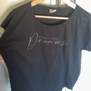 Svart t-shirt Female Passion S/M - Svart t-shirt från Female Passion med trycket 'Chase your Dreams' i ljus text på framsidan. Modellen är klassisk med rund hals och korta ärmar. Materialet är mjuk bomull som känns skönt mot huden. Perfekt för chill dagar eller när du vill ha en inspirerande vibe. Den har använts även köpt som natttröja, knappast använd
