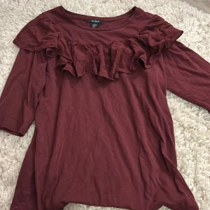 Vinröd topp med volang från Lindex - Supersnygg vinröd topp från Lindex med bred volang längs axlarna och bröstet. Modellen har rund hals och trekvartsärm, perfekt för dig som gillar lite extra detaljer. Mjuk och skön bomull som känns chill och enkel att styla.