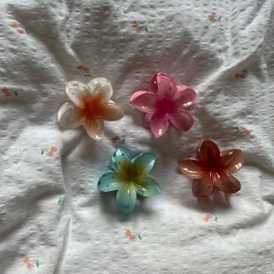 Färgglada blommiga hårklämmor - Fyra superfina blommiga hårklämmor i form av stora plastblommor. Färgerna är vit-orange, rosa, blå-gul och brunröd. Perfekt för att ge din frisyr en somrig och lekfull vibe. Blommorna har glansig finish och är gjorda av plast.