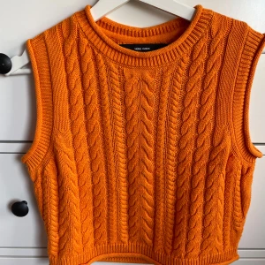 Orange stickad väst från Vero Moda - Säljer en orange stickad väst från Vero Moda i storlek S och är nästan helt oanvänd. Västen har ett klassiskt kabelstickat mönster, ribbade kanter och är ärmlös. Perfekt att styla över en skjorta eller t-shirt för en trendig look.