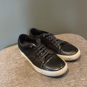 Svarta sneakers från Lanvin med lackad tå - Snygga svarta sneakers från Lanvin med vit sula och cool lackad tå. Skorna har snörning och en klassisk rund tå. Perfekta för dig som gillar stilrena men ändå unika detaljer. Märket syns på sulan och materialet är en mix av skinn och lack.