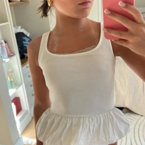 Vit linnetopp med volangkant - Säljer en söt vit linnetopp med breda axelband och volangkant nedtill. Toppen har en enkel, stilren design med en liten spetsdetalj vid halsringningen. Perfekt för varma dagar och enkel att matcha med jeans eller kjol.